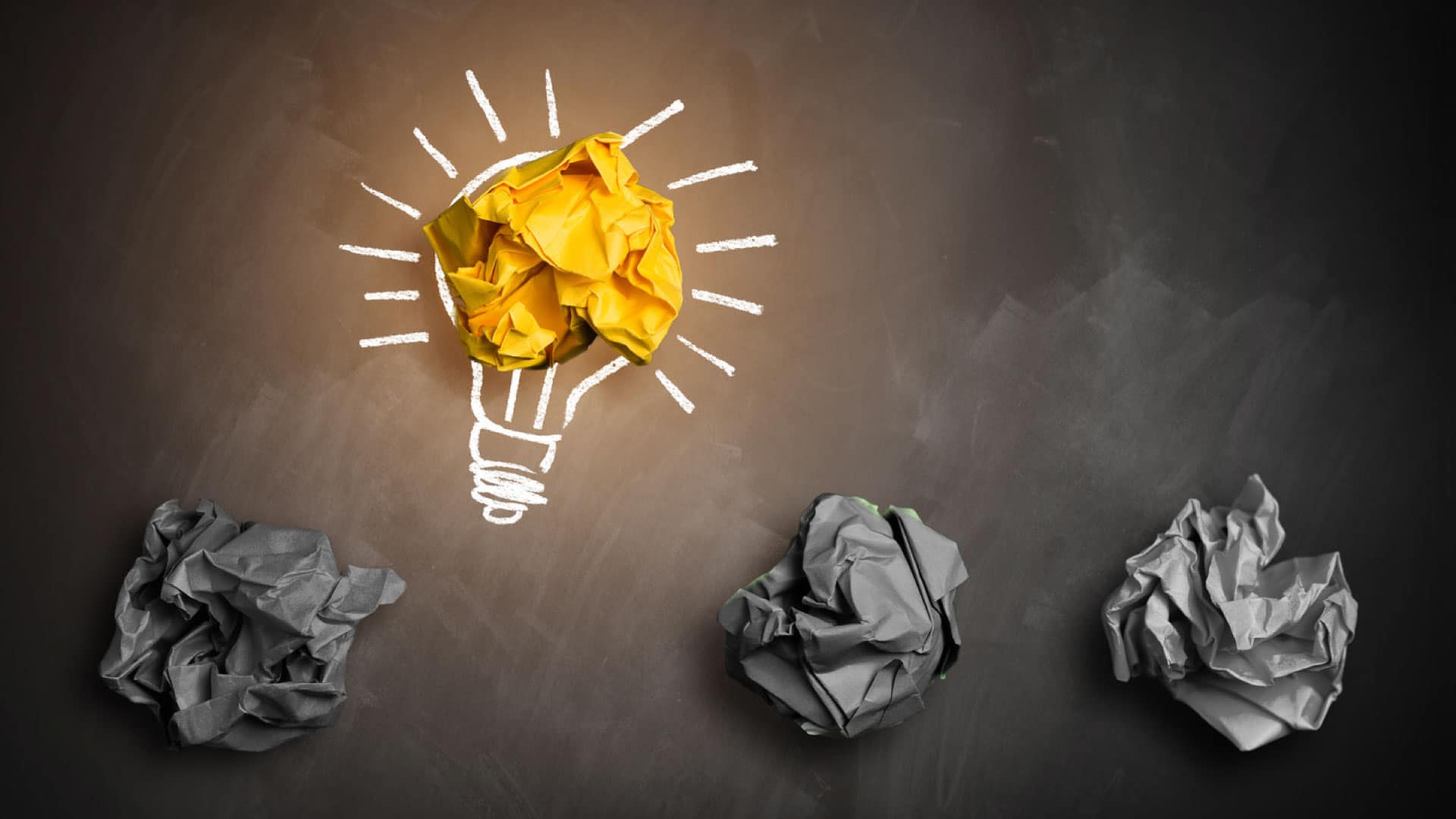 Four crumpled paper balls, three dark and one golden, positioned at the center of a drawn lightbulb on a table, representing Financial Literacy for Career Success.